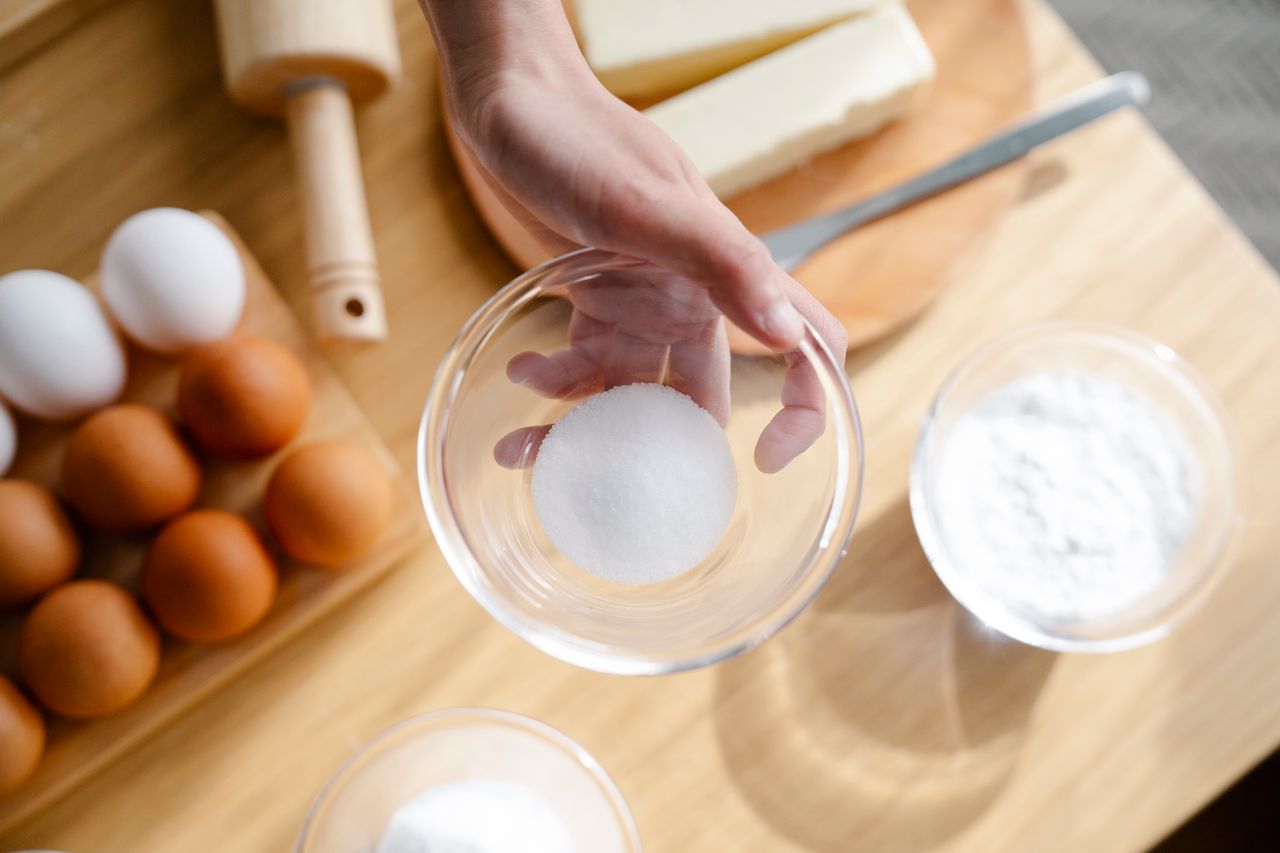 Close-up of hand holding sugar in glass bowl with baking ingredients like eggs and flour.