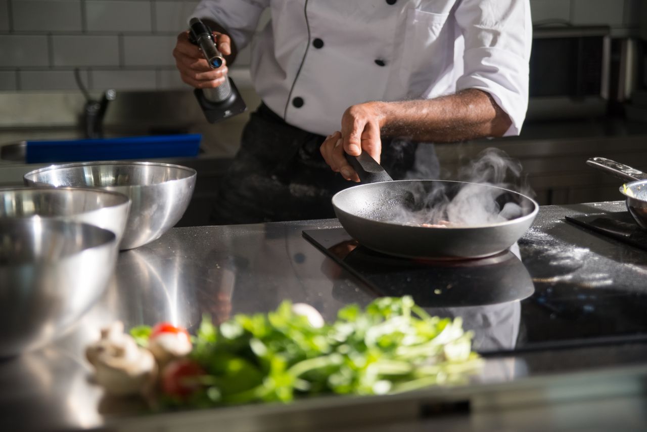 about-us-01 Chef prepares food in a sleek kitchen using a frying pan on an induction cooktop.